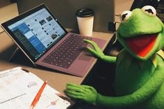 Kermit Grodan sitter vid ett skrivbord med en bärbar dator, kaffe och papper med anteckningar.