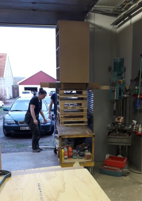 Två personer lyfter en halvpall med material i ett garage med verktyg och en bil i bakgrunden.