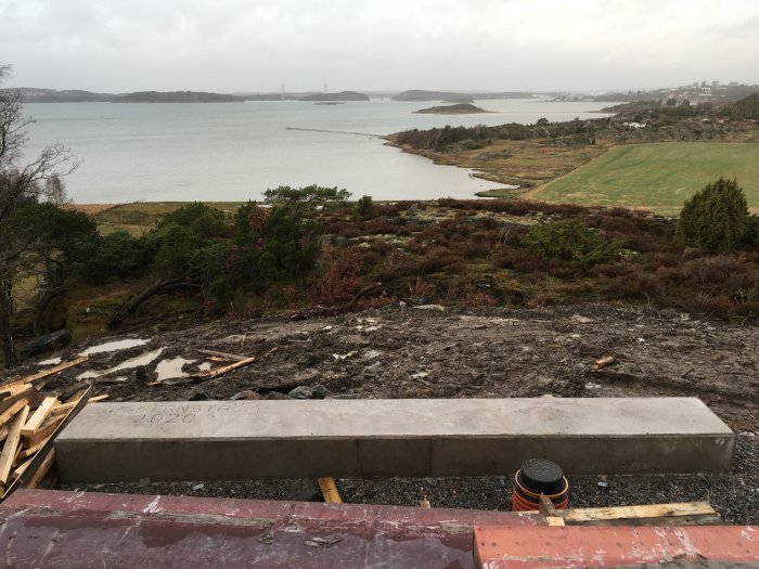 Betongbalk för altanpelare framför en byggarbetsplats med pågående markarbete, utsikt över havet i bakgrunden.