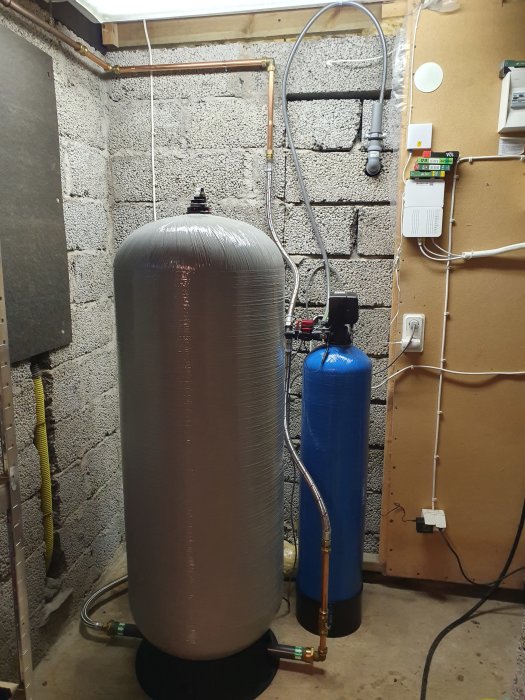 Vattenreningsfilter och hydrofor i källare, med rördragning och isolering synlig.