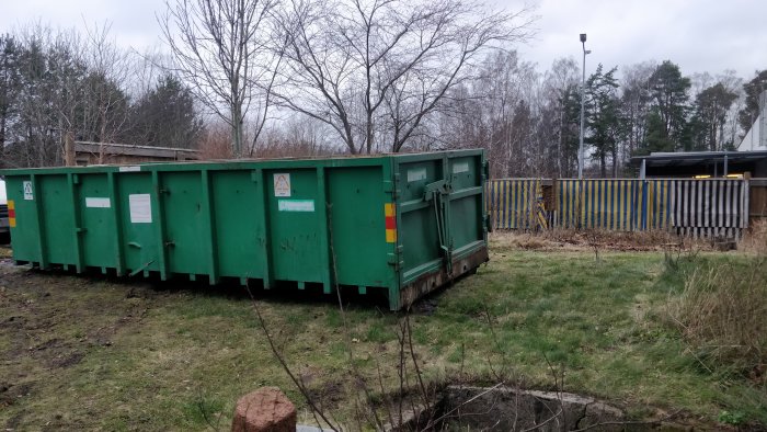 Grön container för byggavfall på ett gräsbevuxet område framför gula byggstaket.