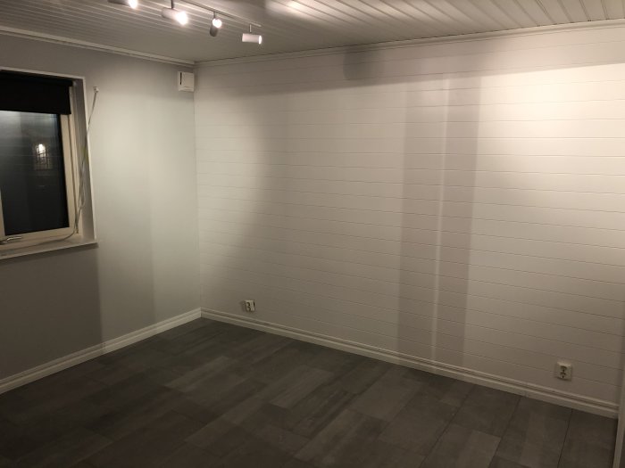 Nylagd panel på sovrumsvägg och grått golv, belysning i taket och fönster till vänster.