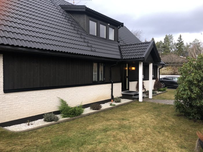 70-talshus med mörk fasad och tak, svart träpanel och vit underdel, med en trädgård framför.