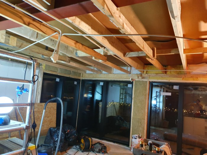 Renoverat tak med nytt träregelverk och isolering, arbetsmaterial syns inuti ett rum med stora fönster mot nattbakgrund.