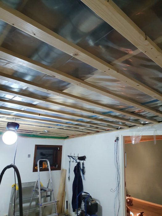Renoveringsarbete med öppet tak, träreglar och plastfolie, verktyg och stege i ett rum.