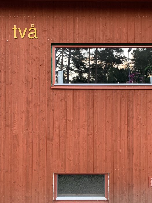 En delvis färdigställd träfasad på ett hus med paneler och ett fönster som reflekterar träd.