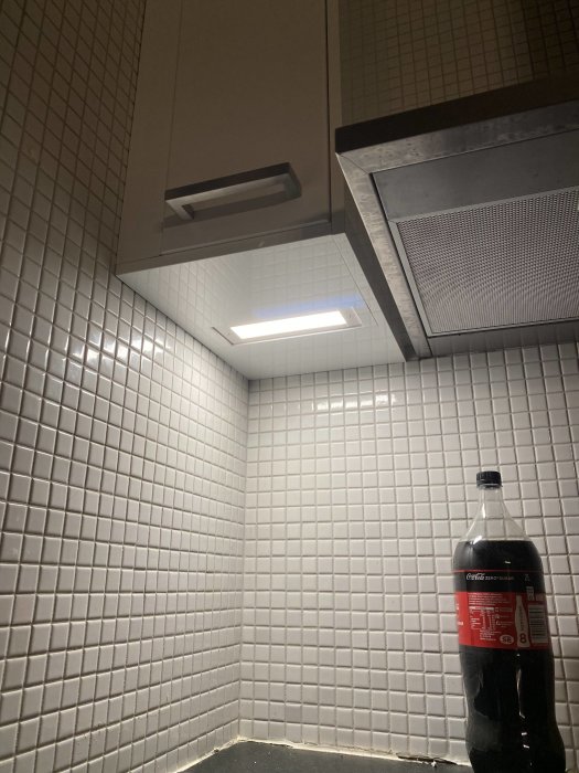 Kökshörna med vit kakelvägg och grå skåpsarg, smalt skåp med 5 cm passbit, Coca-Cola-flaska på bänk.