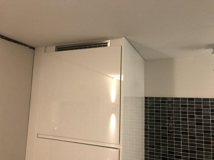 Vitt köksskåp under tak med anslutning till kaklad vägg, synlig ventilationsgrill i hörnet.