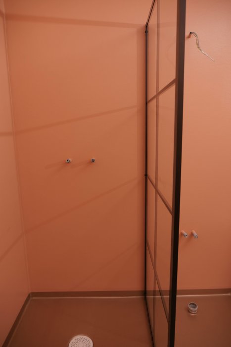 Duschhörna med orange väggar, två synliga rörutgångar för blandare och öppet eluttag.