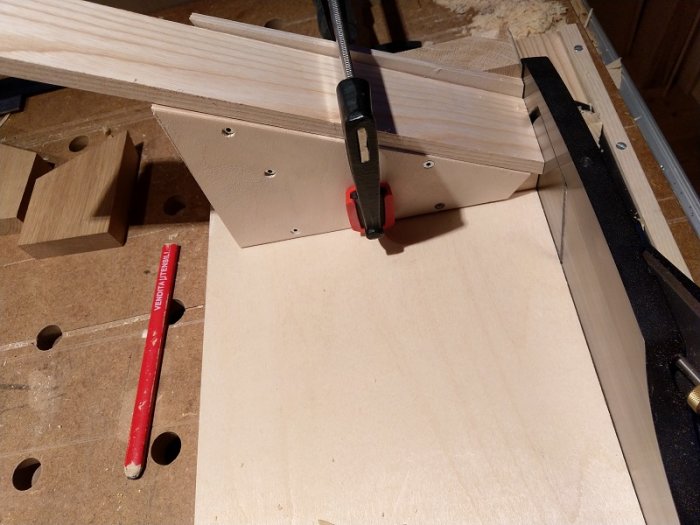 Träplanka fastklämd i stötlåda med extra kloss och snett åsneöra på en arbetsbänk med hål och röd penna.