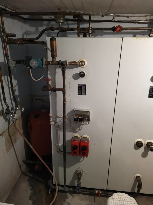 VVS-installation med vit tank, ventiler, rör och mätare i ett pannrum, utloppsrör nere till vänster.