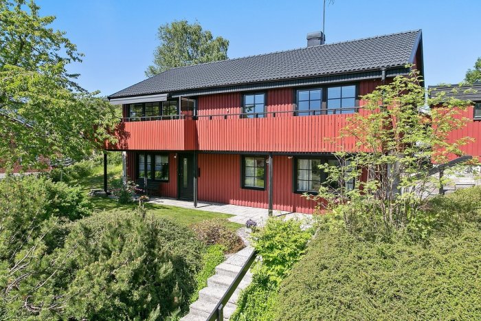 Villa med röd fasad och svarta detaljer, två våningar med balkong, i grönskande omgivning.