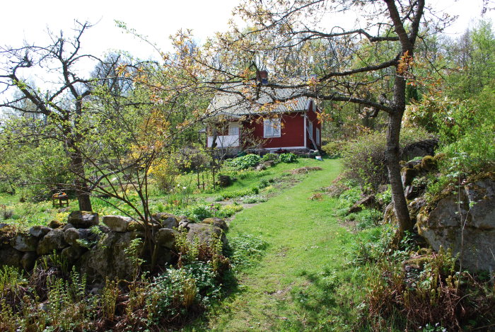 Rödfärgat trähus med två våningar omgivet av en frodig trädgård och stenmur, potentiell plats för tillbyggnad syns till höger.