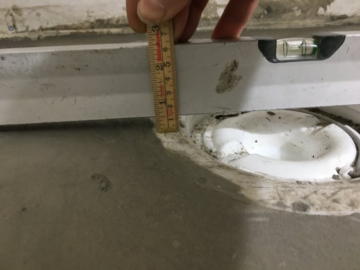 Mätning av golvets lutning nära en dörr med ett vattenpass och tumstock som visar 13mm höjd över en avstånd på 60cm.