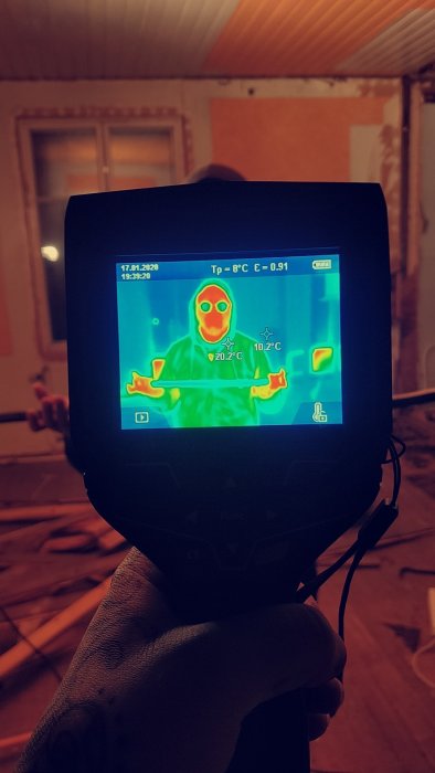 Värmekamerabild av en person hållande ett verktyg i ett rum under renovering, visar temperaturfördelningen.