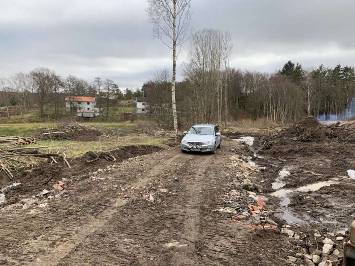 Bil på lerig vägbygge på tomt med högar av jord och spridda byggmaterial i förgrunden.