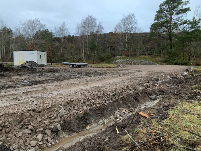 Markarbeten för vägbygge med en arbetsbod och släpvagn på en lertomt med spridda stenblock.
