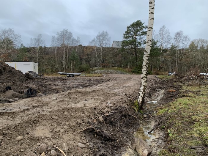 Vägarbete på tomt med synliga hjulspår i leran, arbetsbod och släpvagn i bakgrunden.
