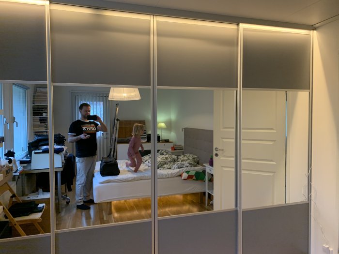 Rörigt sovrum reflekterat i en spegelgarderob med vuxen och barn, lådor och oöppnade flyttlådor.