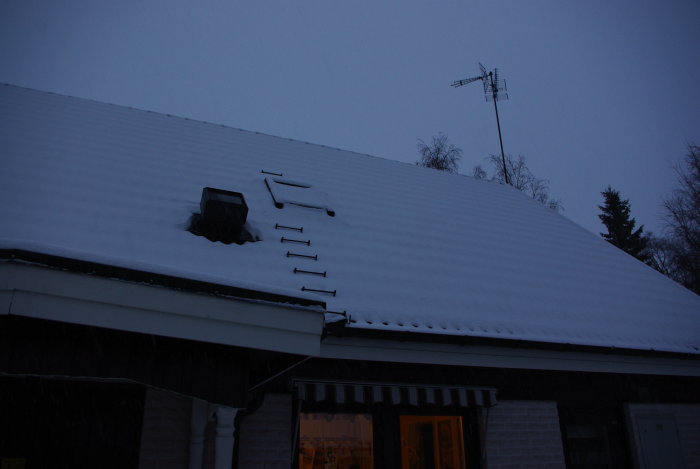 Snötäckt hustak med takfönster och antenn under vinterkvällen.