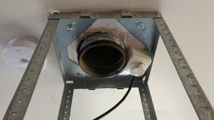 Höjd ventilationsfläkt med synlig anslutningsrör och spår av tejp, intill rökdetektor på en vägg.