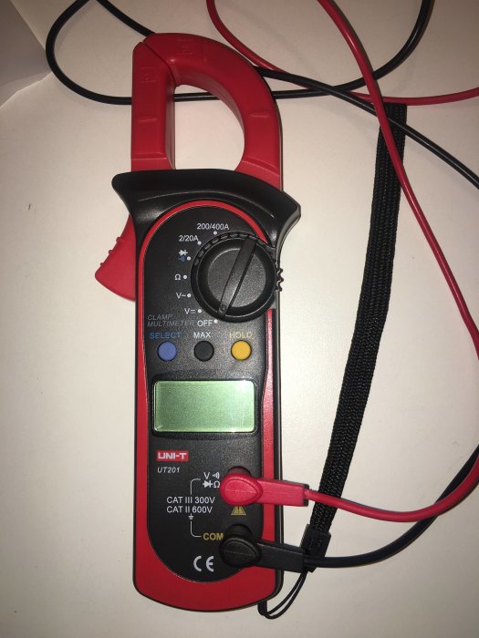 En UniT tångamperemätare med röda och svarta mätledningar och en grön display, ligger på ett vitt underlag.