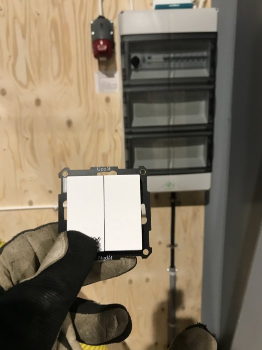 Hand i handske som installerar en smart belysningskontroll bredvid ett elskåp och rörskyddade kablar på en plywoodvägg.