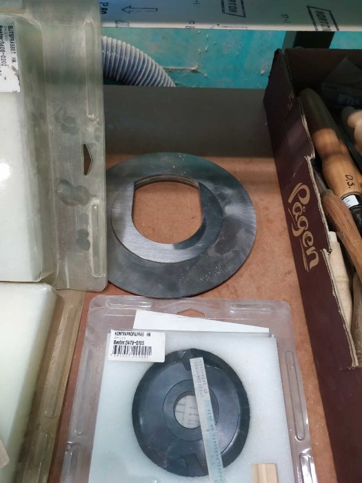 Olika byggmaterial och verktyg, inklusive en rund metallbricka, på en rörigt organiserad arbetsplats.