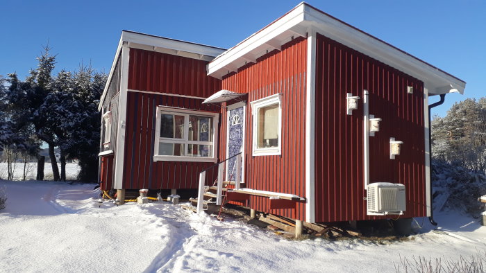 Röd fritidsstuga med luftvärmepump i snötäckt landskap, omgjord från förråd till permanent boende.