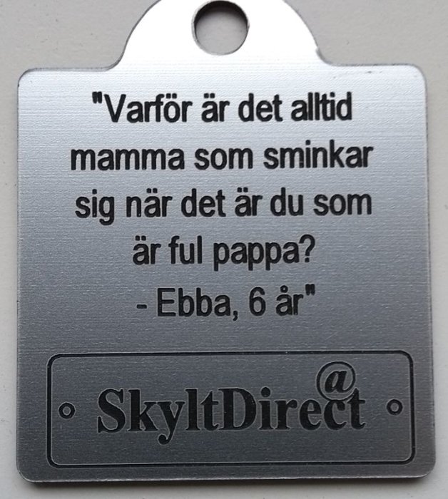 En metallskylt med citatet "Varför är det alltid mamma som sminkar sig när det är du som är ful pappa? - Ebba, 6 år" och logotypen SkyltDirect.