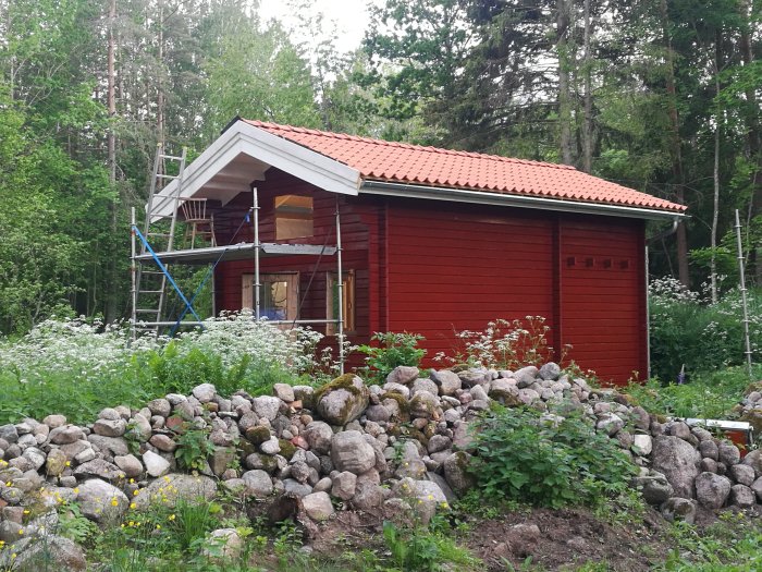 Nyomålat rött trähus med byggnadsställning, omgivet av en stenmur och grönska.