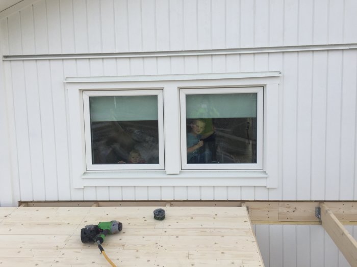 Under uppbyggnad av garagetak med synliga reglar och verktyg, barn tittar ut genom husets fönster.