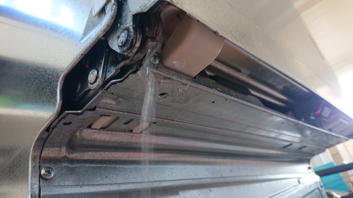 Vatten läcker från en diskmaskins underkant nära gångjärnen och en vit plastdetalj.