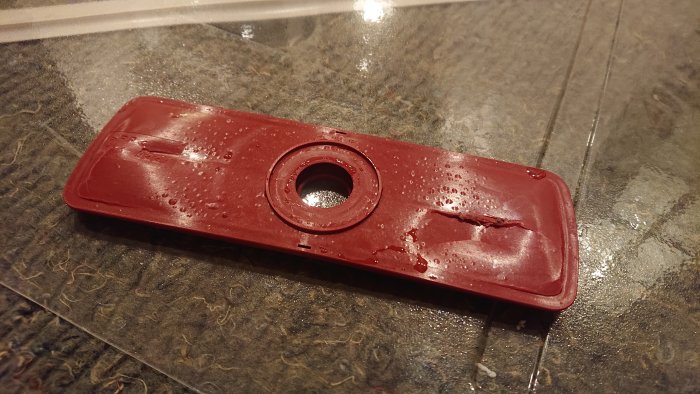 Röd sprucken plastdel från diskmaskin med vattendroppar, ligger på en stenbänk.