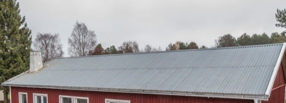 Ett plåttak på en byggnad med röda väggar under en grå himmel, osäkerhet kring taktypen uttrycks.