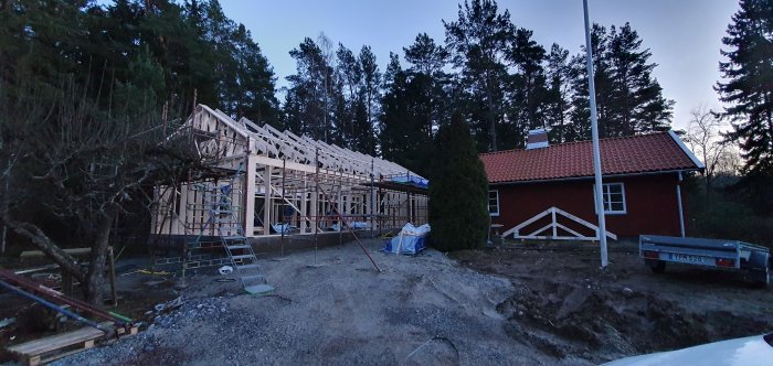 Tillbyggnadsstomme vid röd stuga vid skymning, oordnat byggarbetsplats med takstolar på plats.