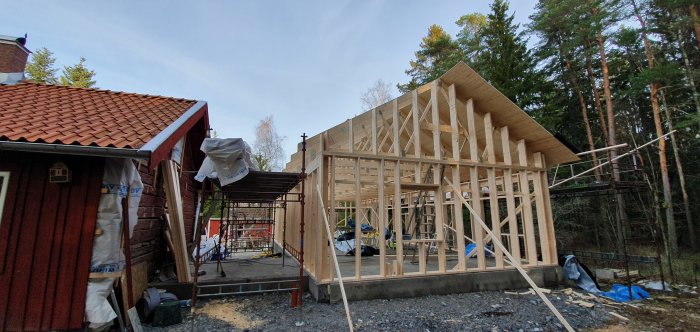 Regnstomme under uppbyggnad bredvid ett äldre rött hus, inramat av skog och med byggmaterial synligt på marken.