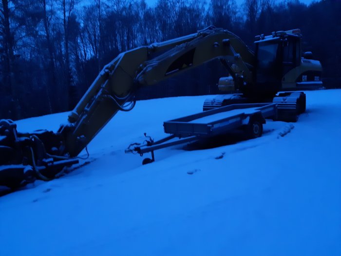 En obrukad grävmaskin i CAT parkerad på snötäckt mark med skog i bakgrunden under skymningen.