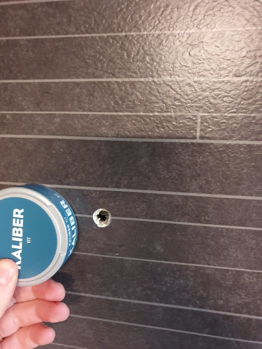 Hand som håller en burk fästmassa framför ett hål i en brun kaklad vägg där ett handdukställ lossnat.