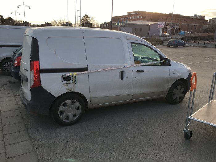 Vit skåpbil med trasig bakdörr säkrad med orange snörstump på en parkering.