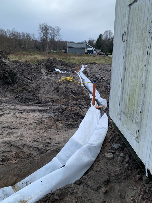 Dräneringsarbete med markduk och rör längs grävd dike vid byggplats.