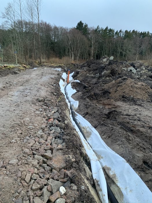 Nyligen grävt dike med markduk och nyinstallerade dräneringsrör vid sidan av en byggplats med skogsbakgrund.