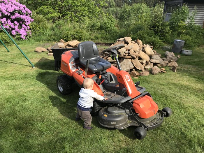 Barn hjälper till att köra en åkgräsklippare på en gräsmatta med en hög av stenar i bakgrunden.