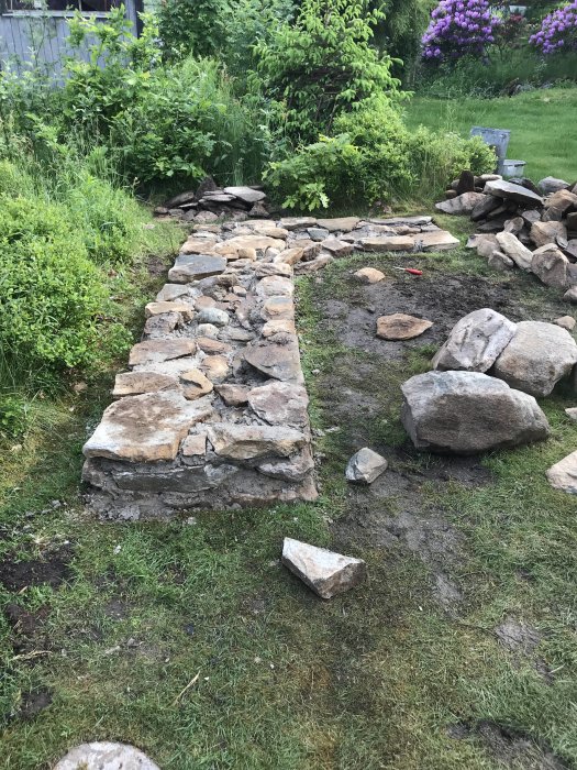 En påbörjad stenkonstruktion för grill i en trädgård med gräsmatta och stenar utspridda runtomkring.