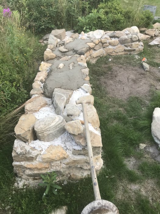 Stenmur i byggnation för grill och rök med trädgårdsspade och omgivande grönska.