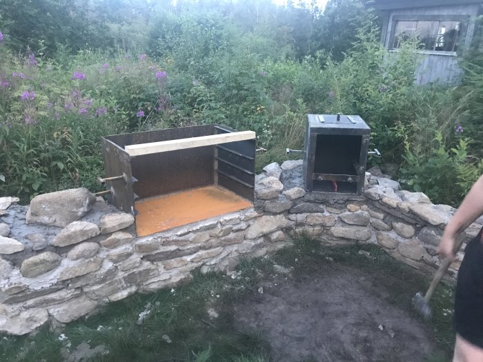 Hembyggd grill och rök i framsidan av ett hus med stenmur och cortenplåtar i skymningen.