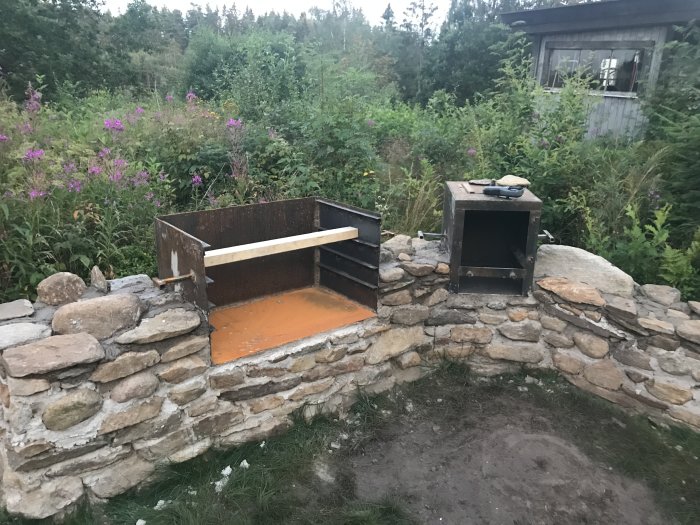 Hjälpreda med stenmurad grill och rökenhet i trädgården under konstruktion, med natur och skog i bakgrunden.