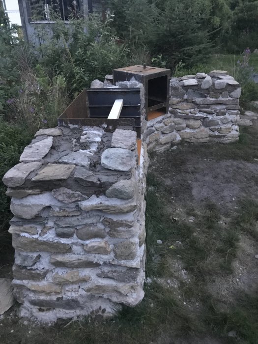 Hemmagjord utomhusgrill av sten med integrerad röklåda under konstruktion i en trädgård vid skymningen.