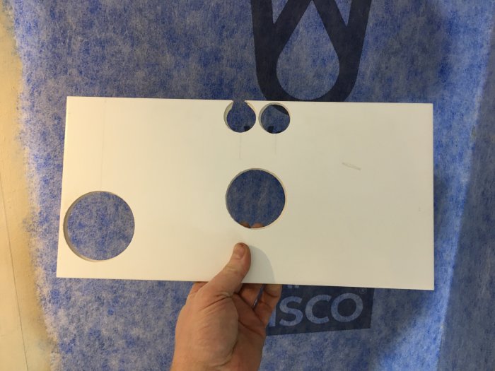 En hand håller upp ett vitt ark med utskurna hål i olika storlekar mot en blå matta.