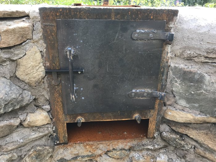Självmurad grill med svetsad svart cortenplåtsdörr inmurad i en stenhög.